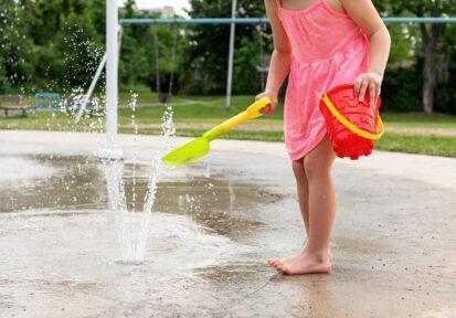 Little girl playing at water splash pad fountain in the park playground during hot summer day.
