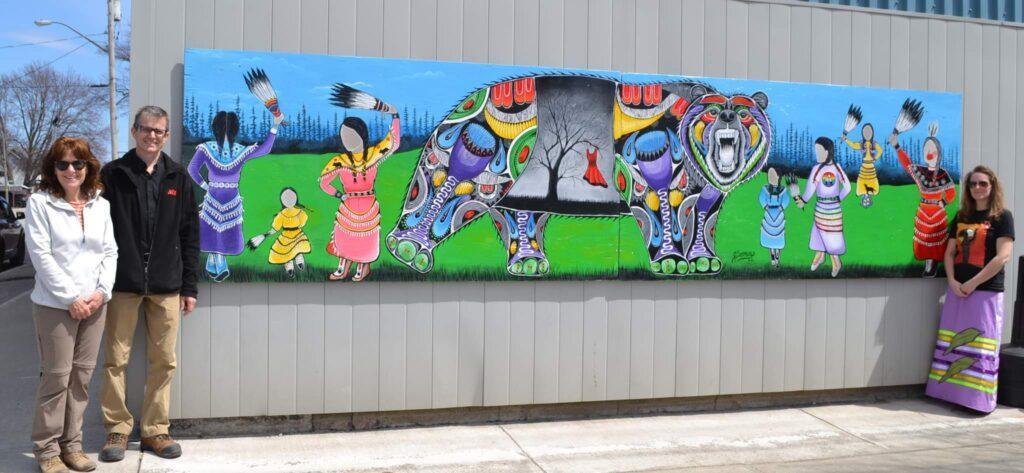 Artist and community members stand proudly in front of colourful mural showing traditional indigenous art.