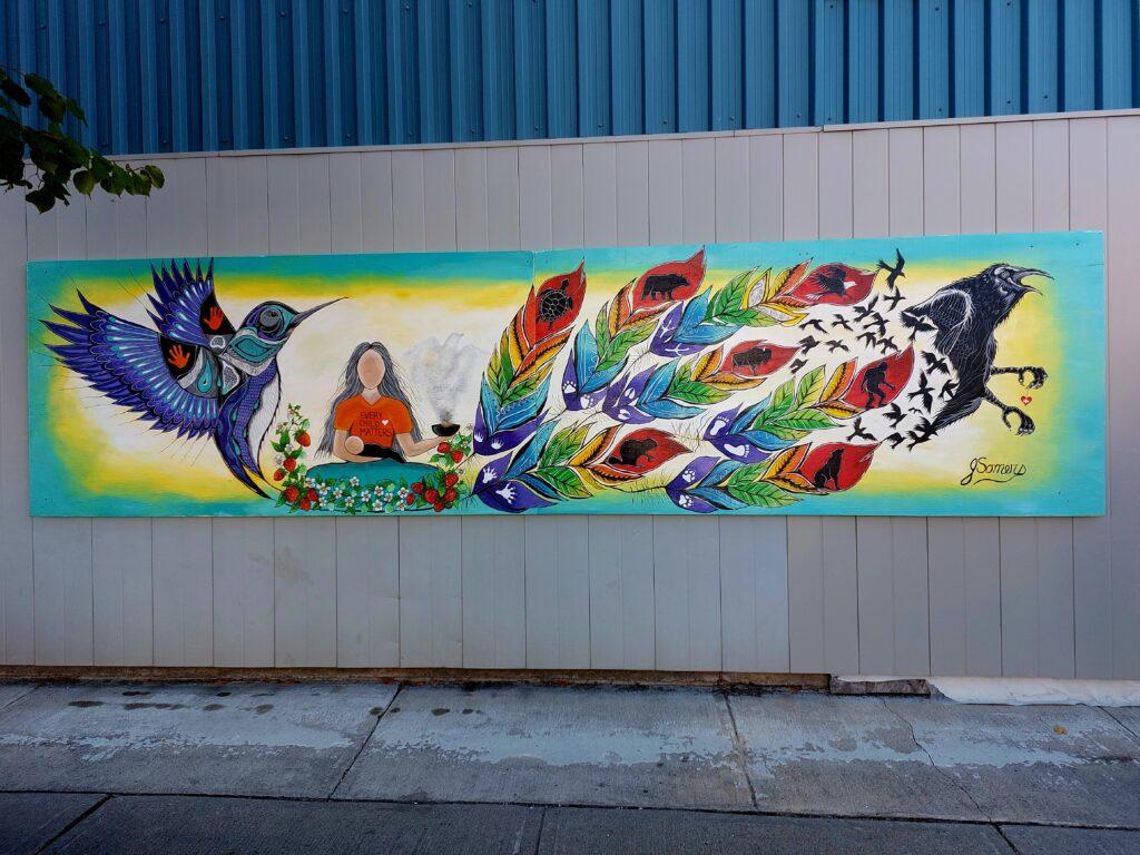 Mural showcases a person sitting between a hummingbird and crow with multicoloured feathers.